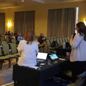 Amanda and Kristina at the table during their presentation. Amanda is speaking through a microphone. 